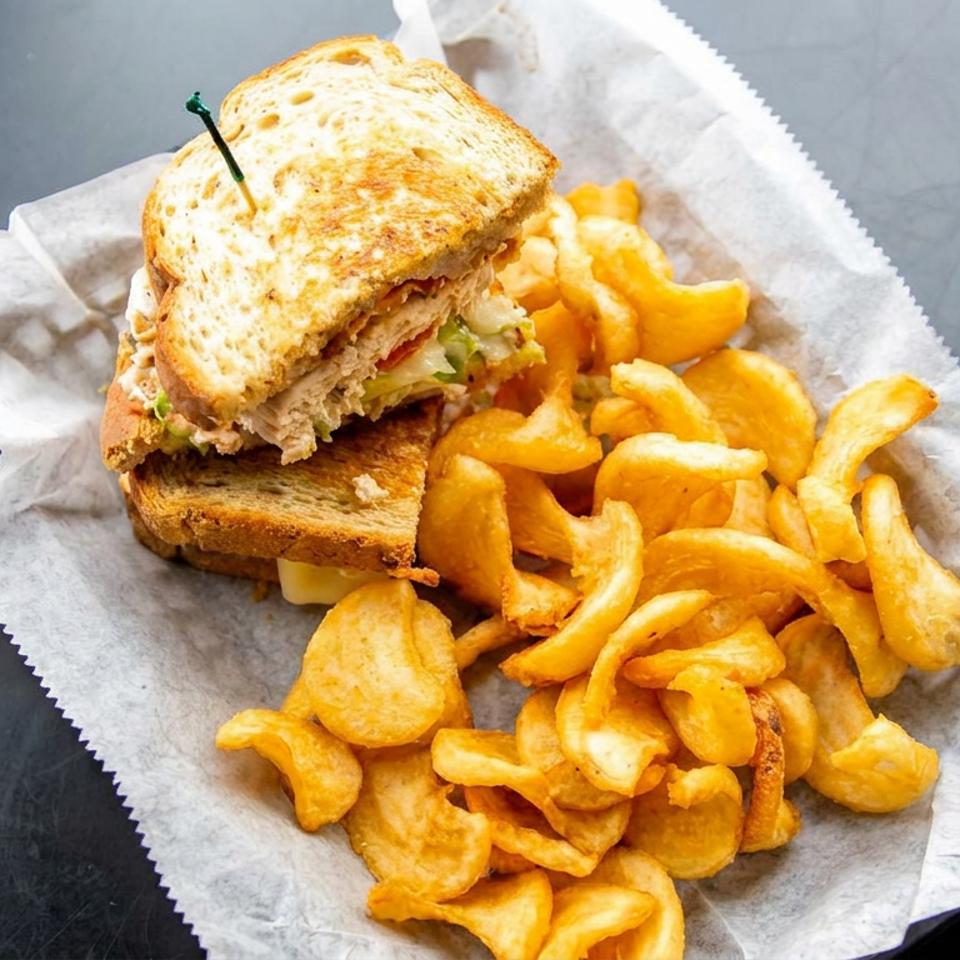 Toasted chicken sandwich with lettuce and tomato served alongside golden kettle chips on parchment paper.