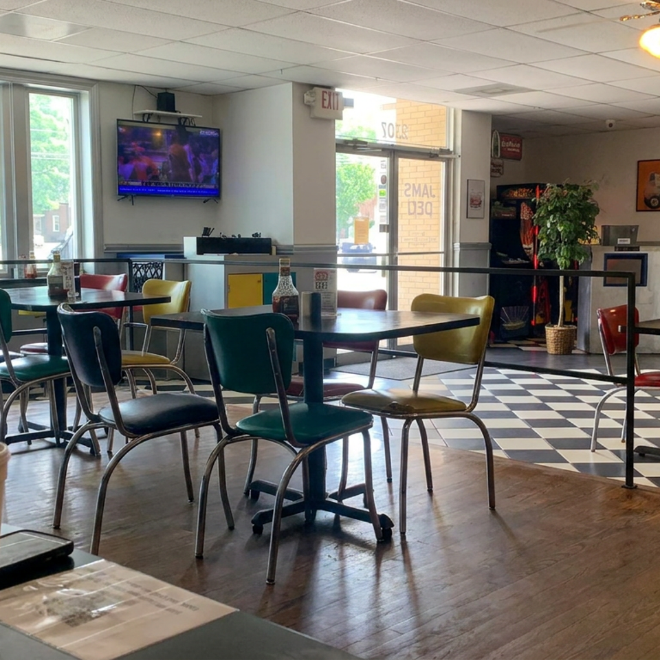 Bright retro diner dining room with colorful chairs, checkerboard floor, television, plants, and tables near large windows.