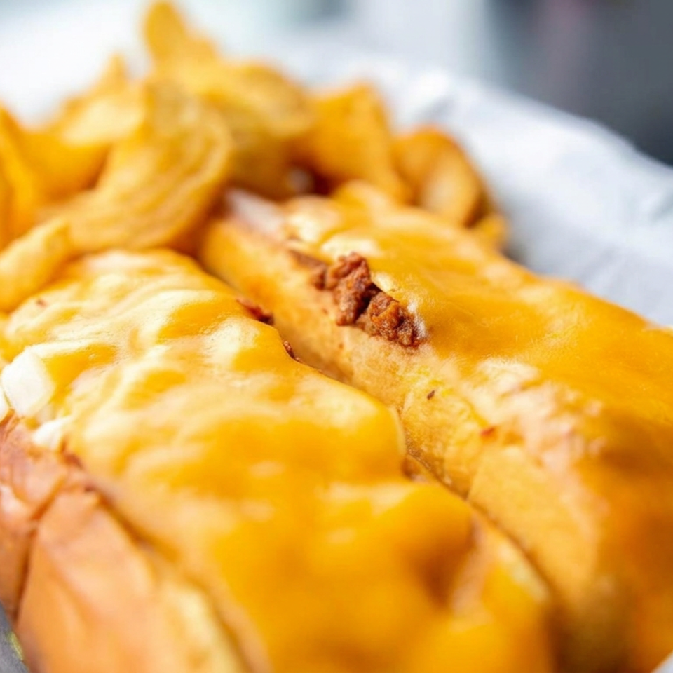 Two chili cheese hot dogs topped with melted cheddar and bacon, served with golden fries in a paper-lined basket.